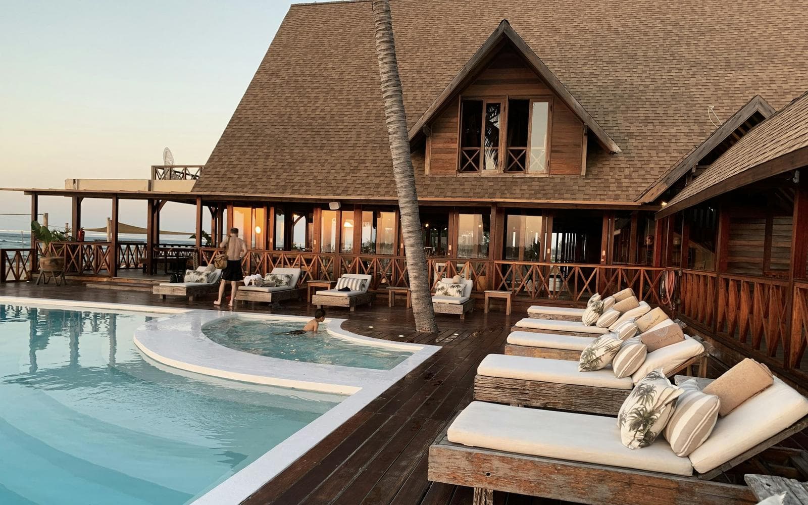 A resort pool lined with palm trees, loungers, and umbrellas beside white hotel buildings.